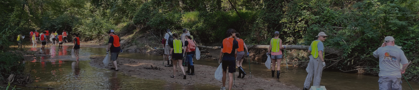 People are walking ina creek wearing bright colored safety vests and holding bags of trash.